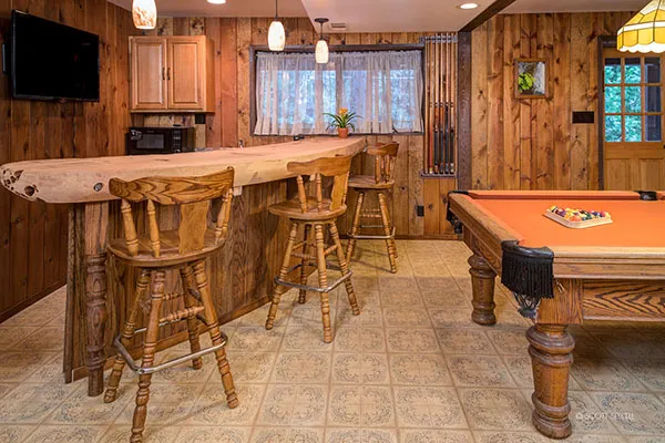 Rustic game room with a wood-slab bar, three wooden barstools, a wall-mounted TV, and an orange pool table.